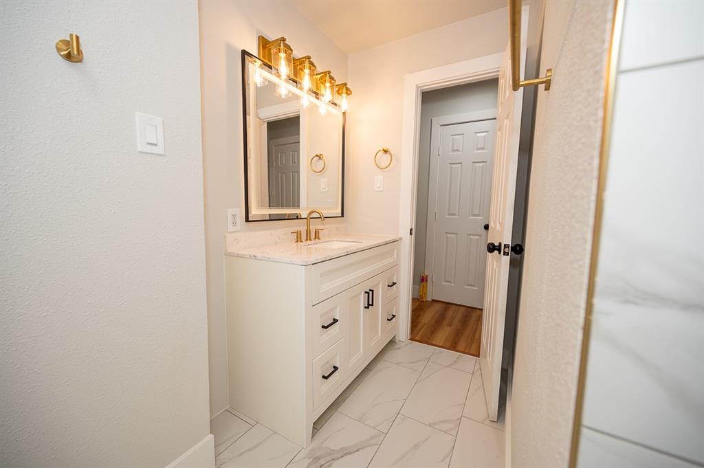 White vanity cabinet with stone-composite countertop, rectangular undermount sink, brass-finish faucet, four-light vanity fixture, and large-format tile flooring