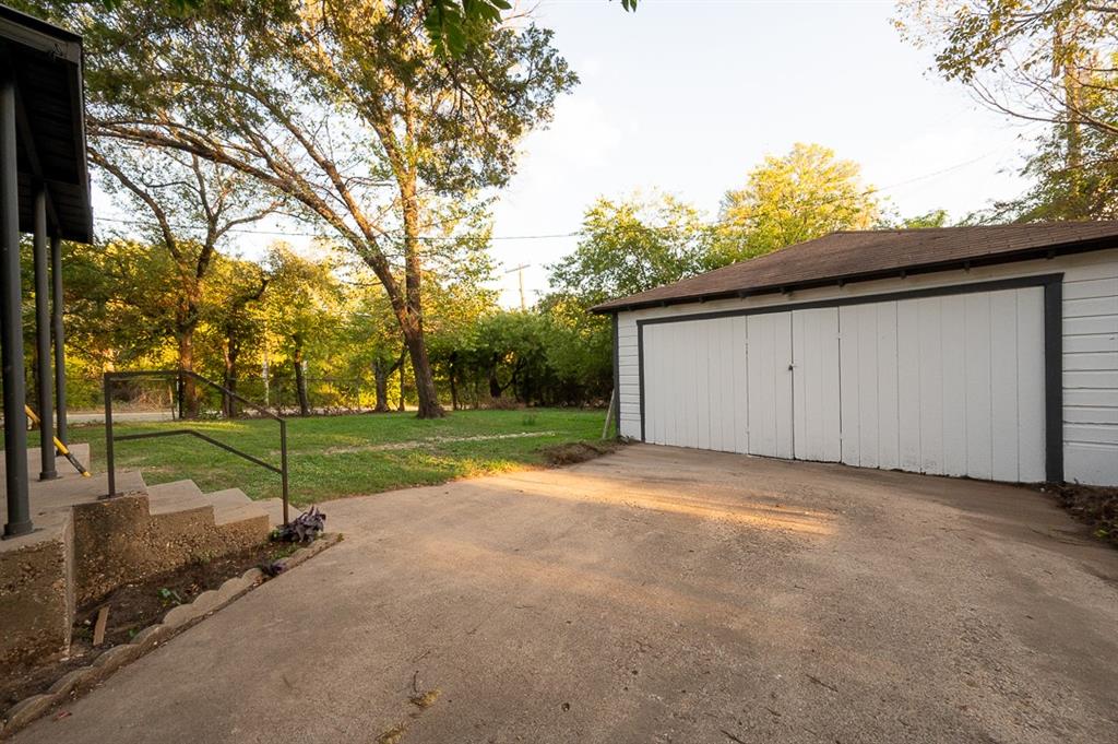 Detached two-car garage with white siding, concrete driveway, concrete stairs with metal railings, and mature trees