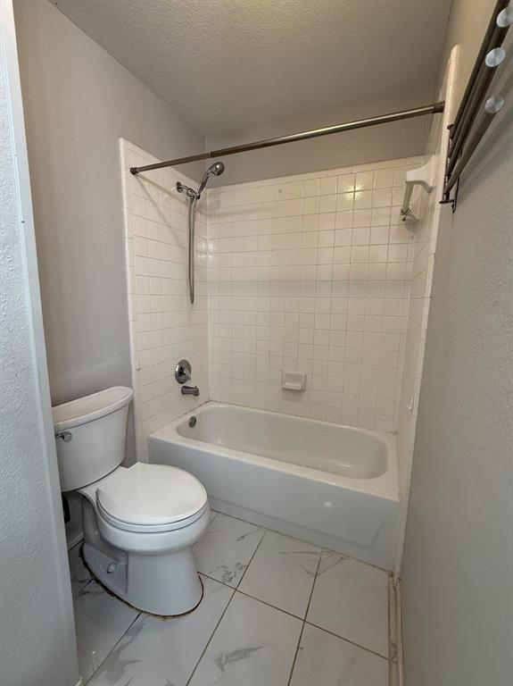 Bathtub and shower combination with white square tile surround, chrome hand-held shower fixture, porcelain toilet, and large-format white tile flooring with grey veining