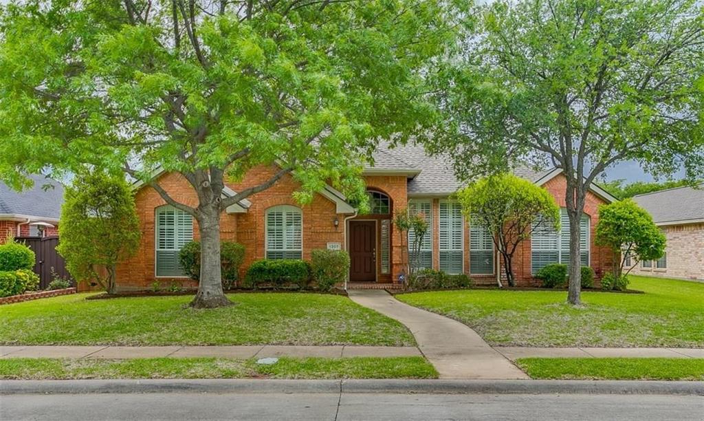 Single-story brick exterior featuring an arched entryway, shingle roof, multi-pane windows with shutters, and mature landscaping