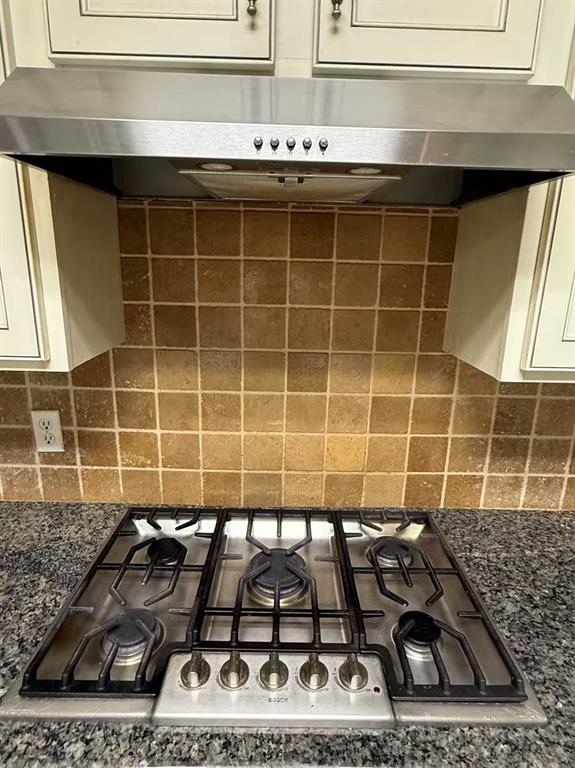 Five-burner stainless steel gas cooktop with heavy-duty grates, matching stainless steel range hood, natural stone tile backsplash, and dark stone-surfaced countertop with light-toned cabinetry