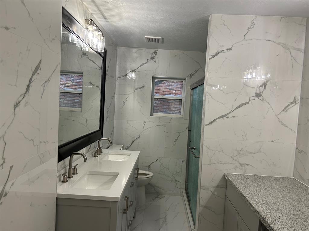 Bathroom featuring floor-to-ceiling large-format veined tile, double-sink vanity with a solid surface countertop, framed wall mirror, and glass-enclosed shower