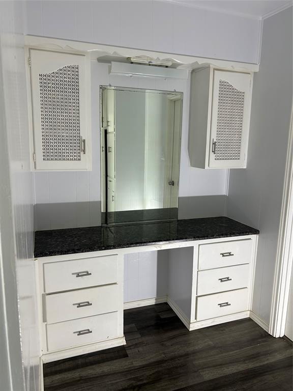 Built-in vanity featuring white cabinetry, dark stone-finish countertop, decorative cabinet door inserts, wall-mounted mirror, and wood-finish flooring