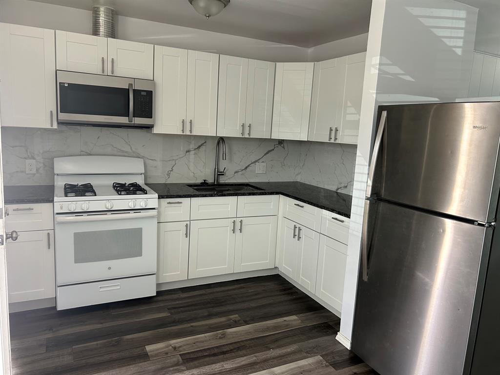 Kitchen featuring white shaker-style cabinetry, dark stone countertops, a full-height stone-pattern backsplash, stainless steel appliances, and wood-finish flooring