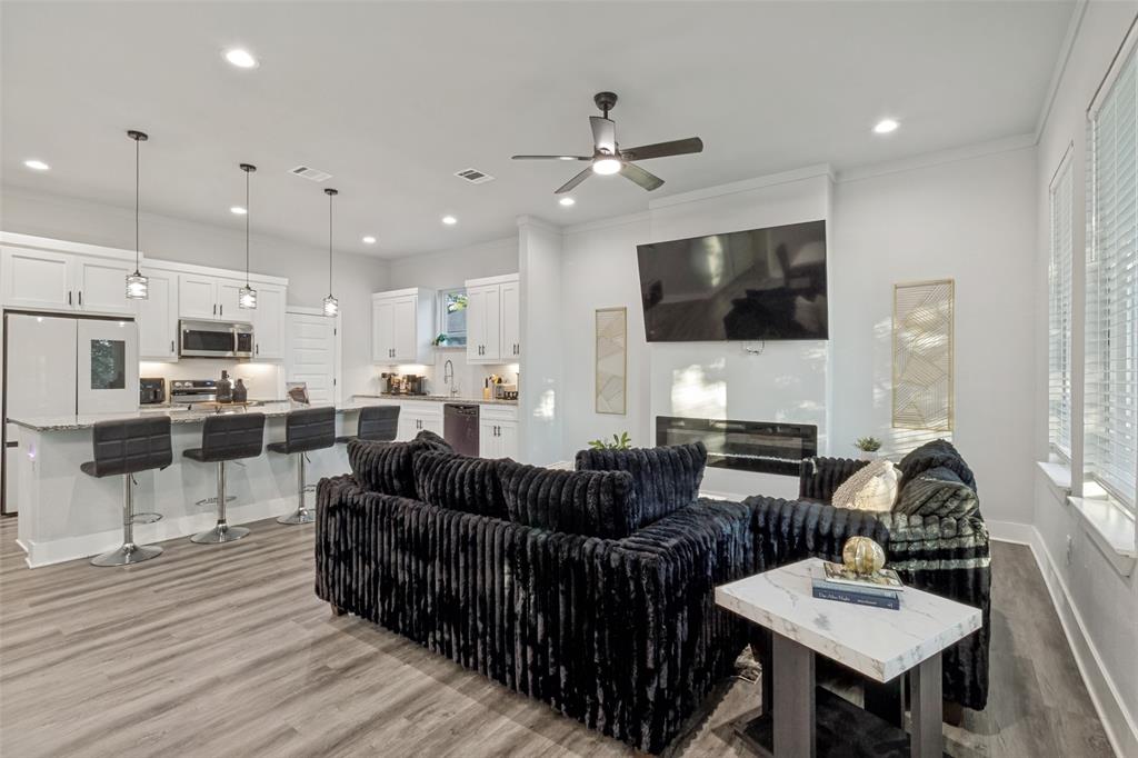 Open concept kitchen featuring white shaker-style cabinetry, an expansive kitchen island with a neutral countertop, built-in stainless steel appliances, recessed lighting, and wood-finish flooring throughout, complemented by a floor-to-ceiling fireplace surround