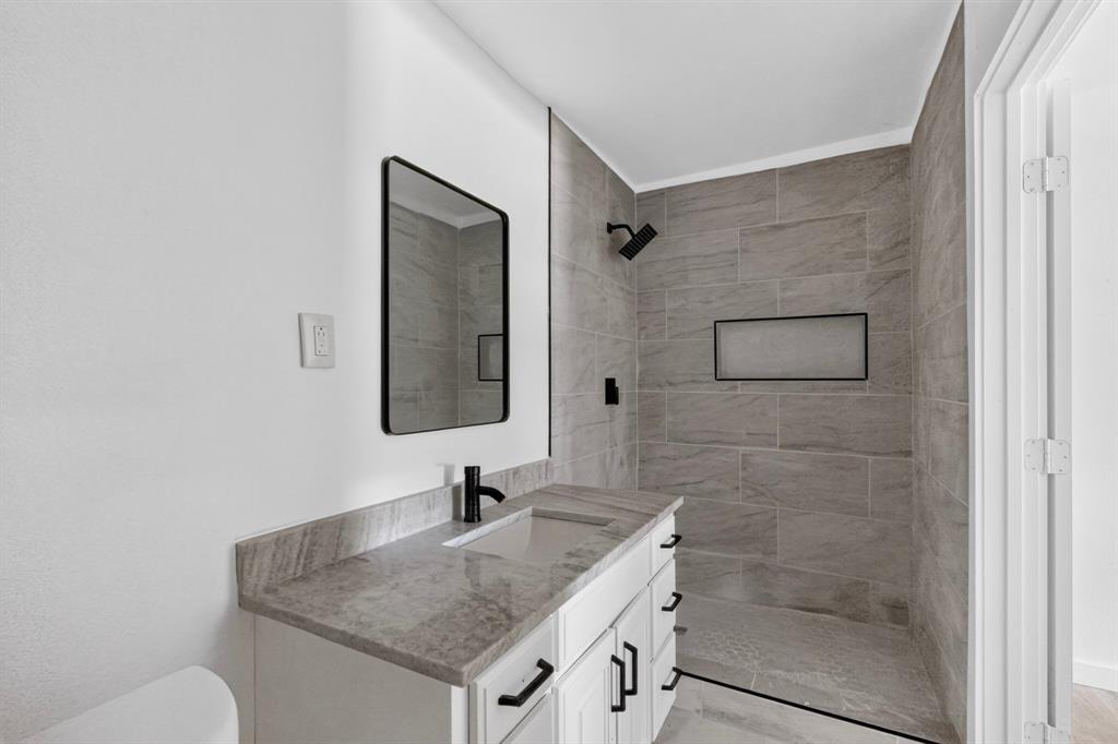 Bathroom featuring a white vanity with stone-finish countertop, undermount sink, matte black hardware, rectangular mirror, and walk-in shower with large-format tile surround and built-in recessed niche