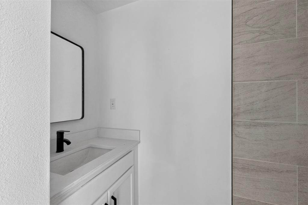 Built-in vanity featuring a solid-surface countertop, rectangular integrated sink, matte black faucet, and rectangular wall-mounted mirror, accented by a textured stone-finish tile wall