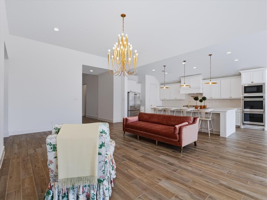 Open-concept interior featuring wood-finish tile flooring, white shaker-style cabinetry, a built-in double oven, quartz-topped kitchen island, and a multi-tiered gold-tone chandelier