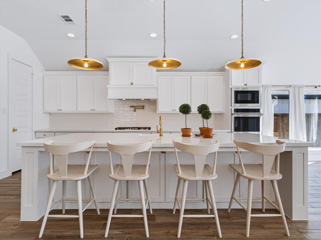 Kitchen featuring an oversized central island with solid-surface countertop, white shaker-style cabinetry, stainless steel built-in double ovens, gold-toned hardware, and three pendant light fixtures