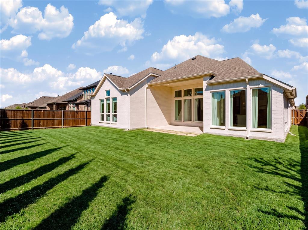 Light-colored brick exterior featuring a covered patio, expansive manicured lawn, full-height multi-pane windows, and wood-style perimeter fencing