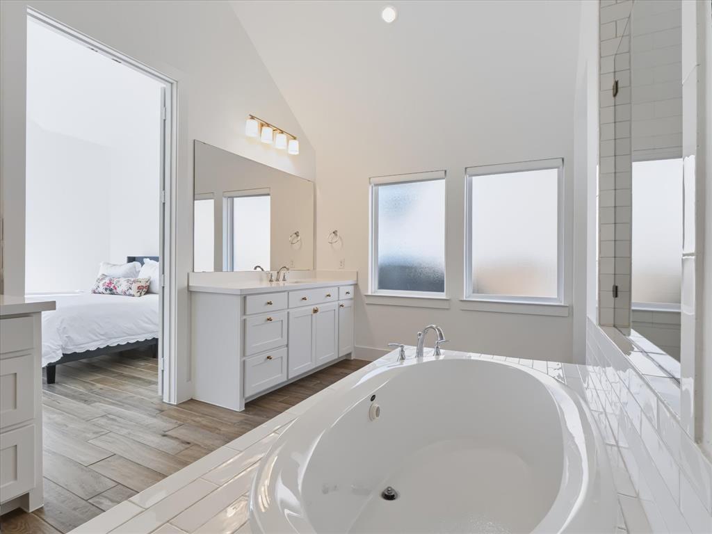 Vaulted ceiling bathroom featuring a white dual-sink vanity, recessed oval soaking tub, subway tile surround, and wood-finish flooring