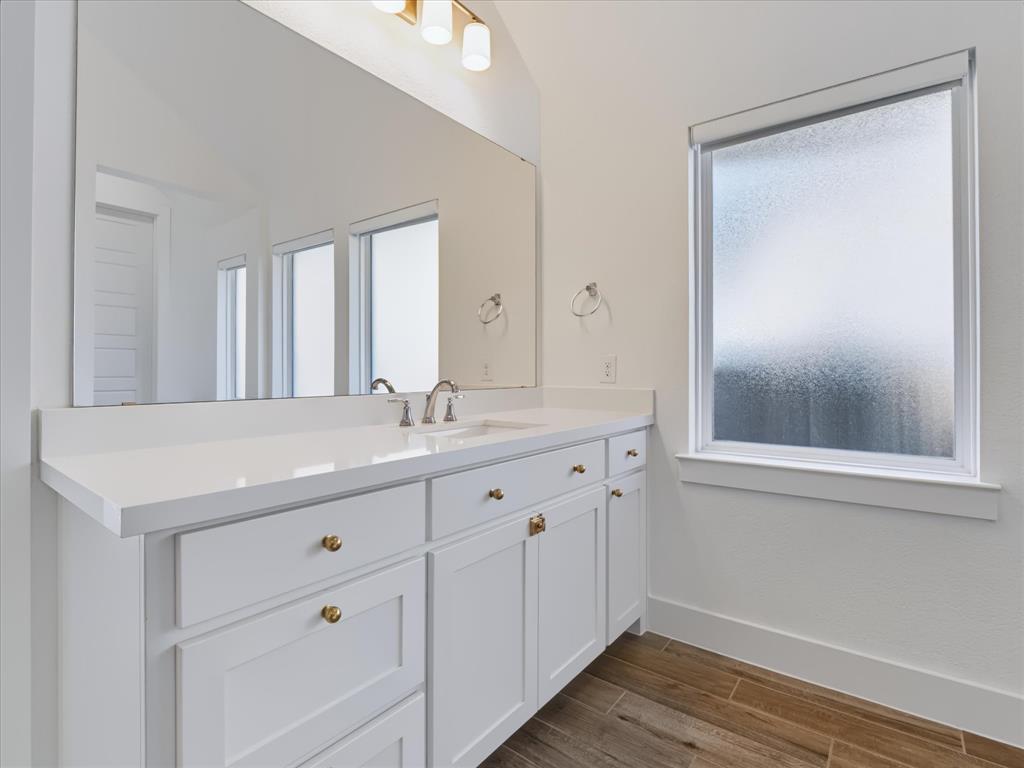 White vanity with a solid-surface countertop, under-mount sink, brushed gold-finish hardware, full-width mirror, and wood-finish flooring
