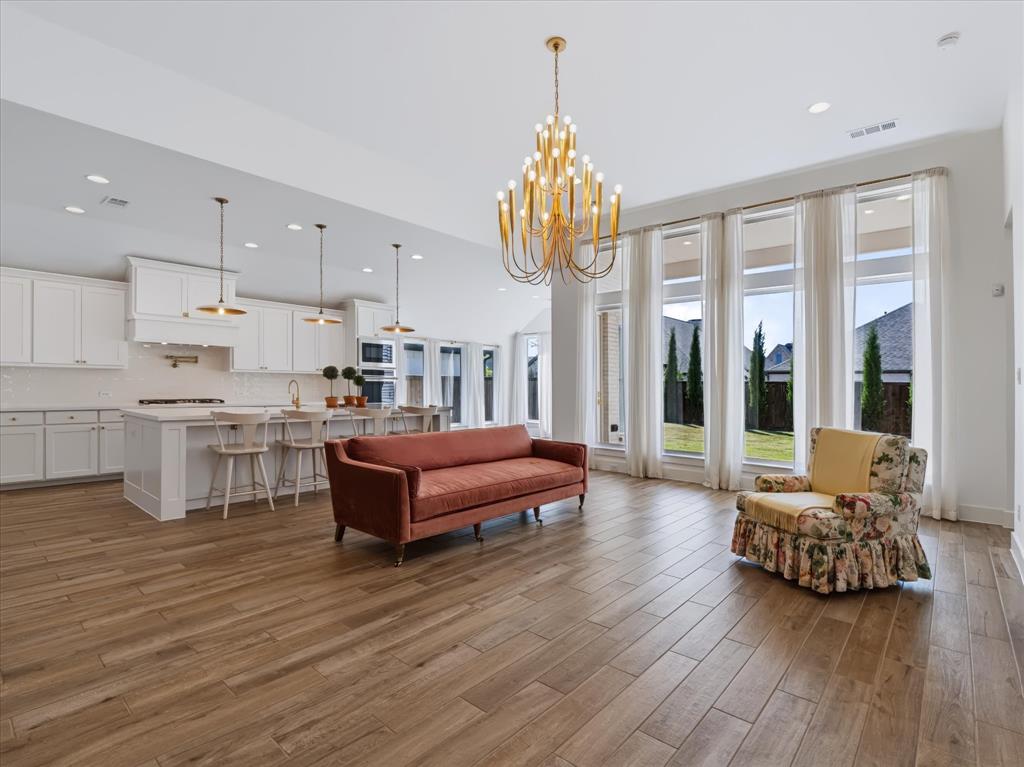 Open-concept interior featuring wood-finish flooring, a kitchen with white cabinetry and a central island, recessed lighting, and a gold-finish tiered chandelier