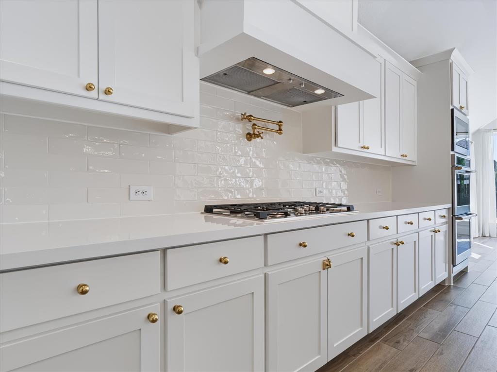 Kitchen featuring white shaker-style cabinetry, a glossy subway tile backsplash, solid-surface countertops, gold-tone hardware, and a wall-mounted pot filler