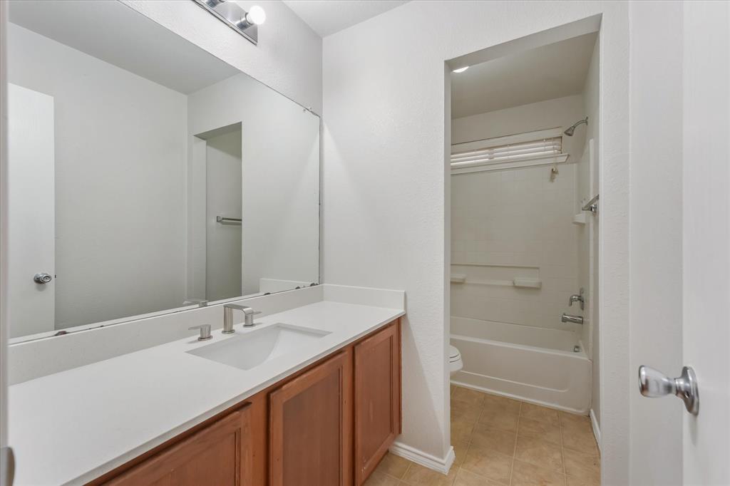 Full-width vanity with a solid-surface countertop, integrated rectangular sink, wood-finish cabinetry, and oversized wall-to-wall mirror, featuring an enclosed tub and shower combination with tile surround Full-width vanity with a solid-surface countertop, integrated rectangular sink, wood-finish cabinetry, and oversized wall-to-wall mirror, featuring an enclosed tub and shower combination with tile surround