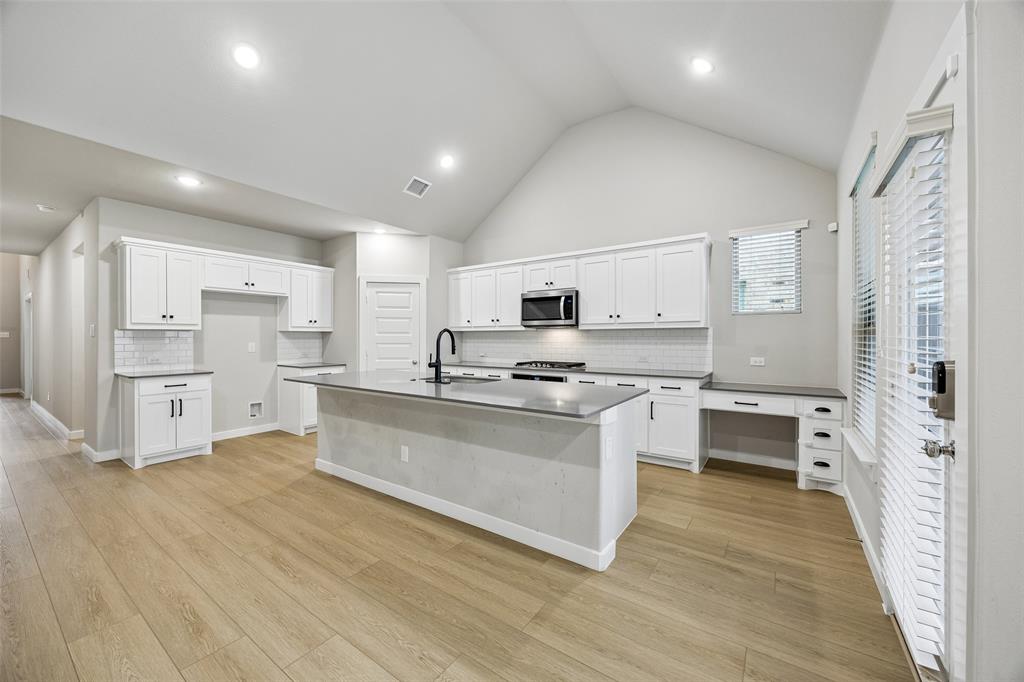 Kitchen featuring vaulted ceilings, a central island with a sink, white shaker-style cabinetry, wood-finish flooring, and a subway tile backsplash