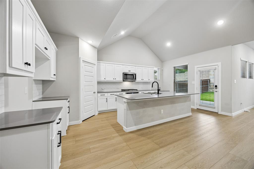 Kitchen featuring vaulted ceilings, wood-finish flooring, a central island with a matte black faucet, and white cabinetry complemented by dark countertops