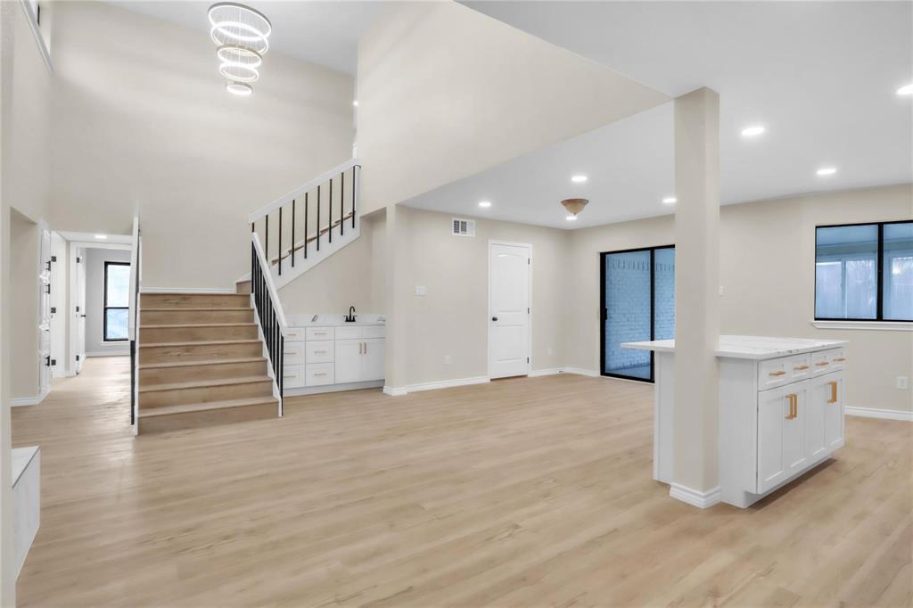 Open-concept interior featuring wood-finish flooring, a double-height ceiling with a multi-ring chandelier, a staircase with matte black metal balusters, and a central island with white cabinetry and quartz-style countertop