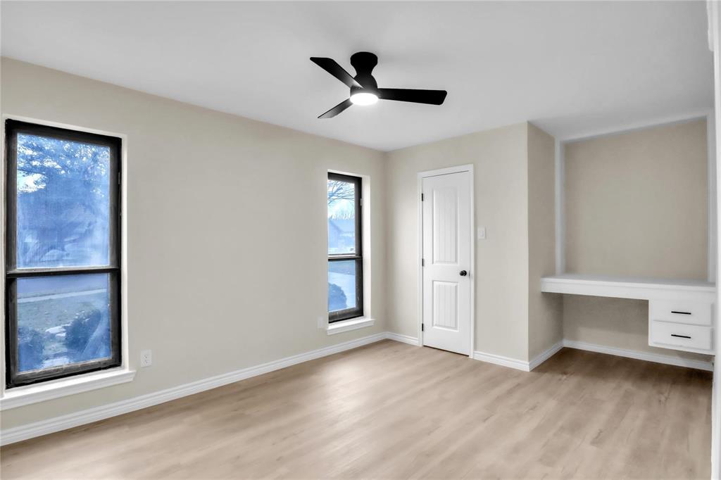 Neutral wall tones with wood-finish flooring, dual windows with black frames, a contemporary ceiling fan with integrated lighting, a solid-core interior door, and a built-in desk with dual storage drawers