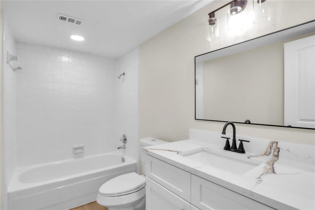 White vanity with stone-patterned countertop, under-mount sink, matte black widespread faucet, rectangular mirror with thin black frame, and tiled tub-shower combination