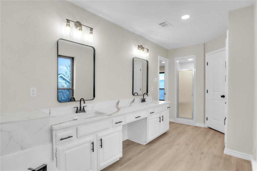 Dual-sink vanity with white cabinetry, light-toned countertop, rectangular wall-mounted mirrors, and matte black fixtures, paired with wood-finish flooring and a walk-in shower enclosure