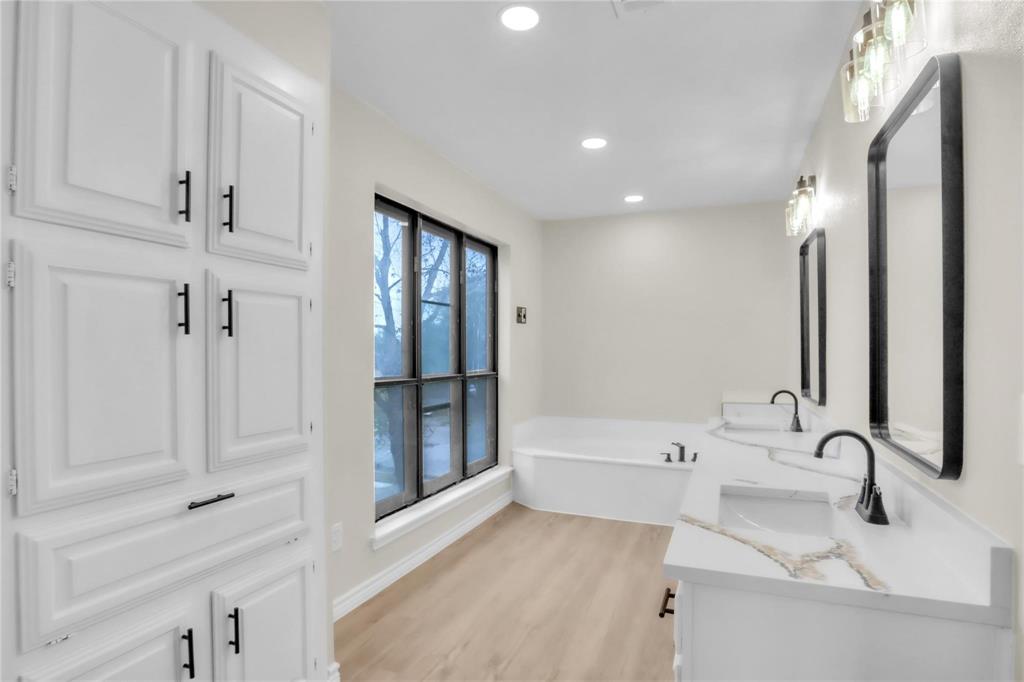 Double vanity featuring white cabinetry, solid-surface countertops, and matte black fixtures, paired with a built-in soaking tub, tall floor-to-ceiling cabinetry, and recessed lighting