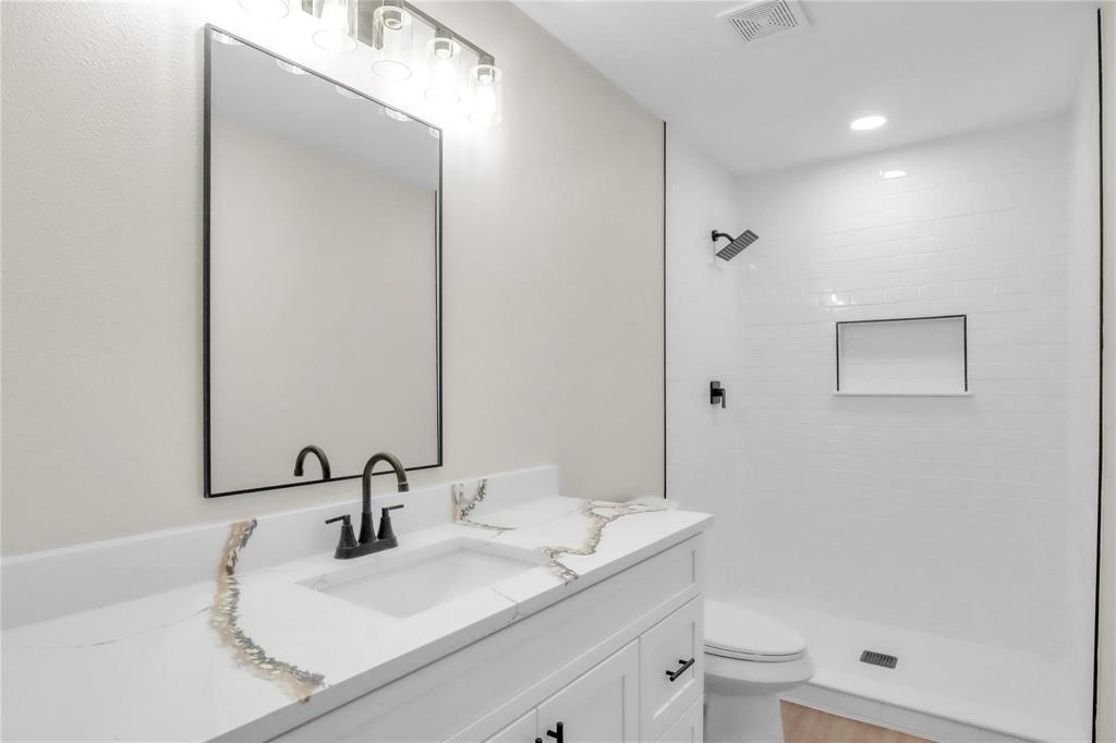 Bathroom featuring a white vanity with a stone countertop, rectangular undermount sink, black metal hardware, framed mirror, and a walk-in shower with white wall tile and a built-in recessed niche