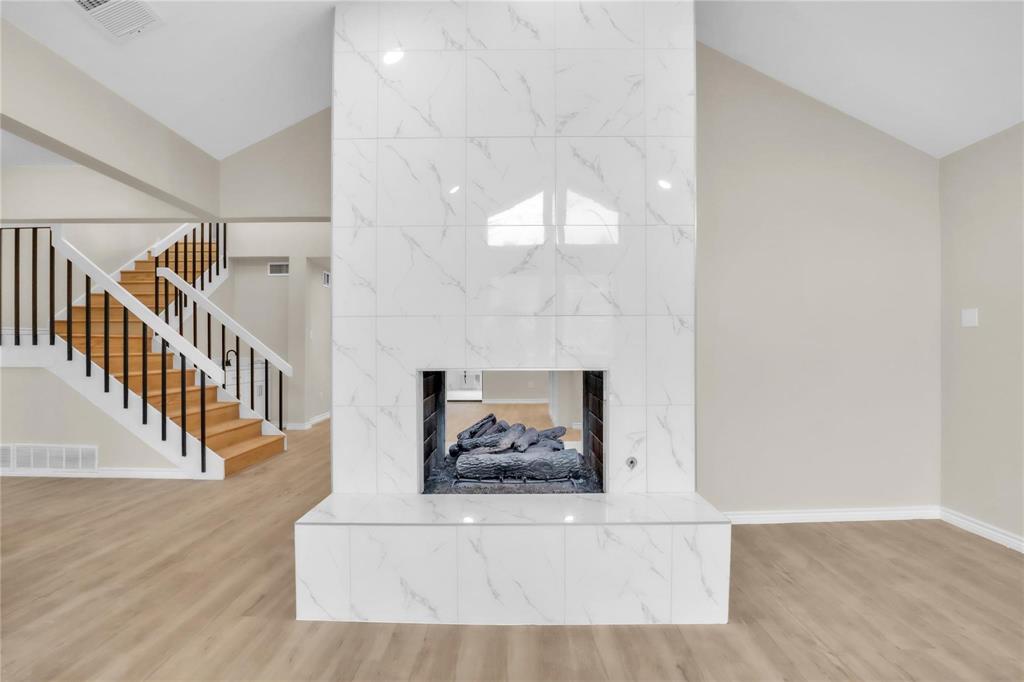 Double-sided fireplace with large-format white stone-finish tile surround and hearth, featuring a staircase with white balusters, black handrail, and wood-finish treads, set against high vaulted ceilings and light wood-finish flooring