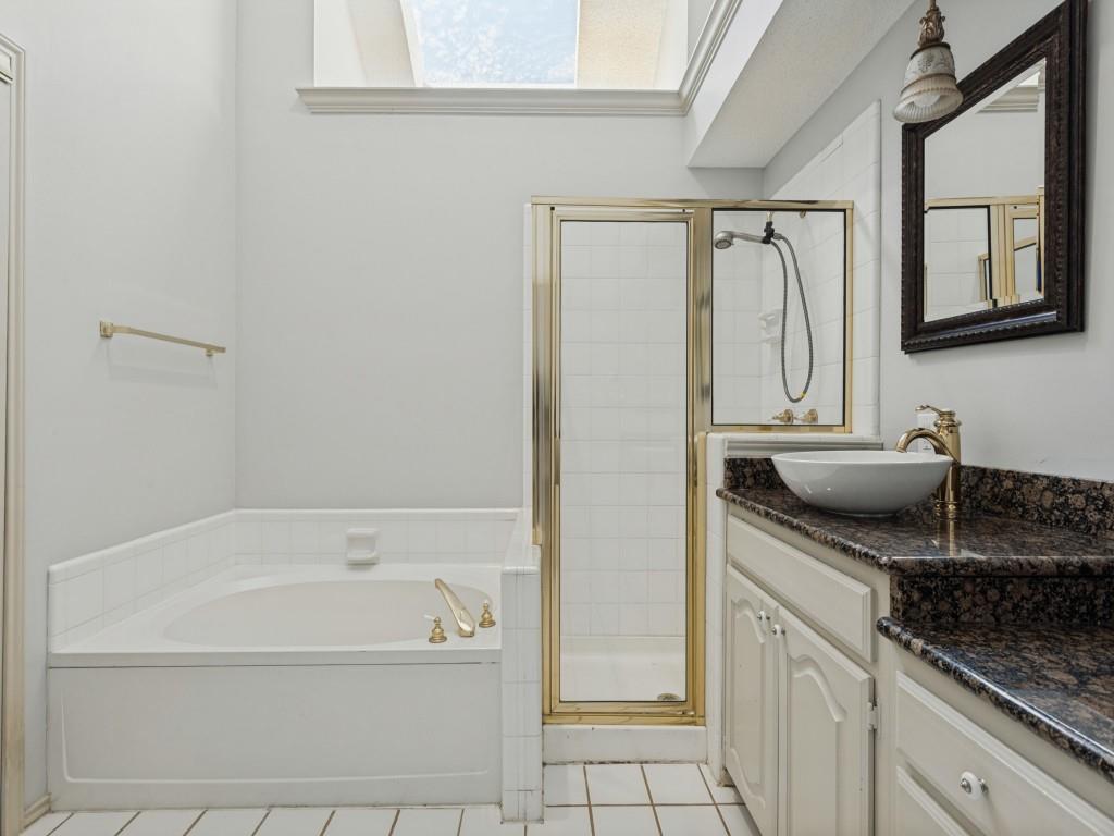 Bathroom featuring a deep soaking tub, glass-enclosed shower with metal framing, vanity with a stone-finish countertop, vessel sink, and overhead skylight Bathroom featuring a deep soaking tub, glass-enclosed shower with metal framing, vanity with a stone-finish countertop, vessel sink, and overhead skylight