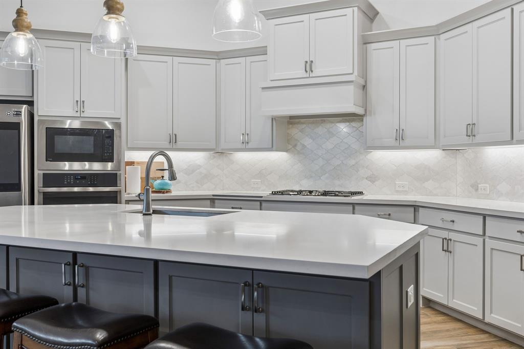 Kitchen featuring light gray shaker-style cabinetry, quartz countertops, a multi-tonal diamond pattern tile backsplash, stainless steel built-in microwave and oven, and a center island with contrasting charcoal-toned cabinetry