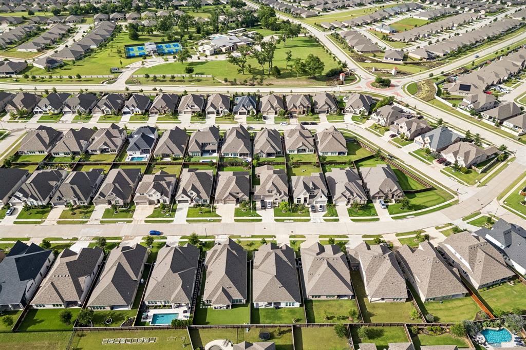 Aerial perspective of a planned community featuring uniform residential roofing, paved street infrastructure, landscaped green space, and a community recreation area with tennis courts