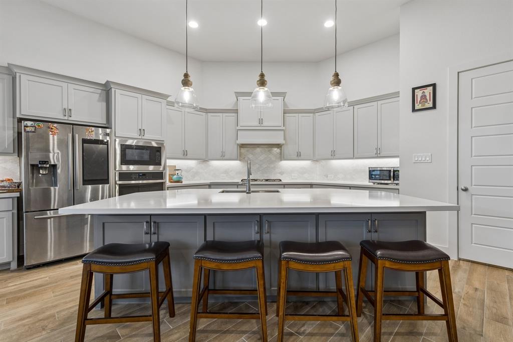 Kitchen featuring a central island with a solid surface countertop, light gray shaker-style cabinetry, stainless steel built-in appliances, herringbone pattern tile backsplash, and three glass pendant lights