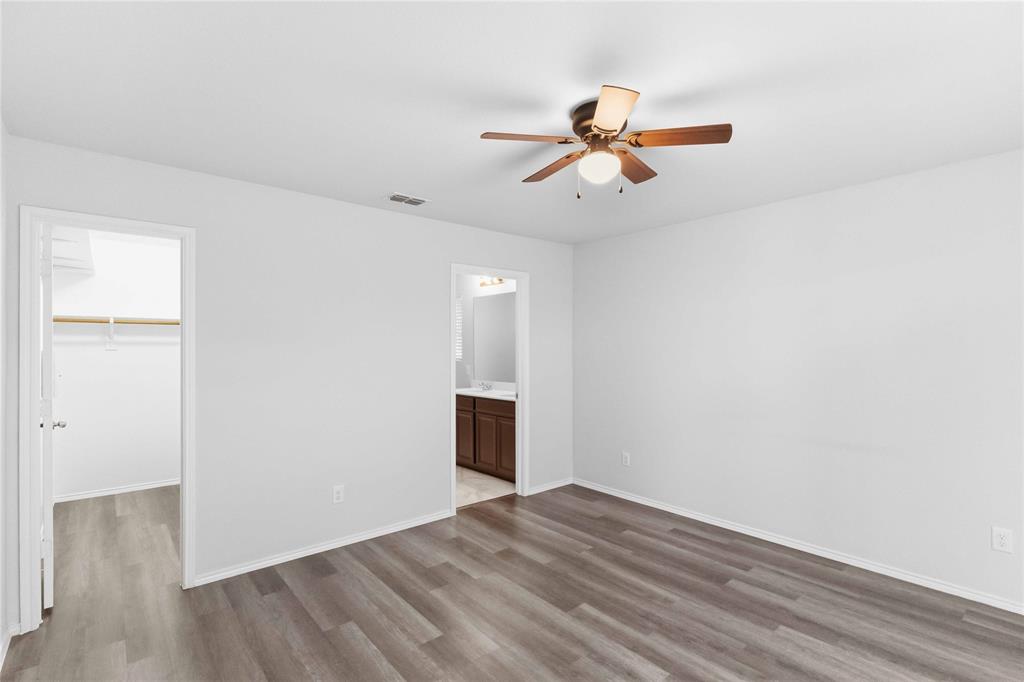 Room featuring wood-finish flooring, a central ceiling fan with integrated lighting, and access to a walk-in closet and a separate bathroom with dark wood-toned cabinetry
