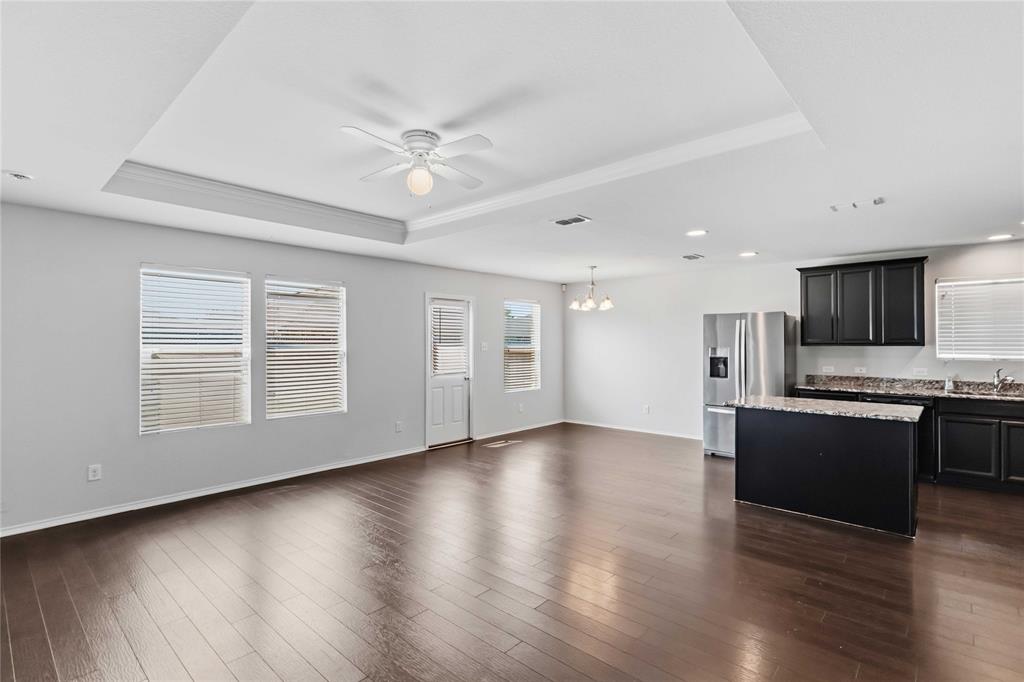 Open-concept interior featuring wood-finish flooring, tray ceiling with integrated lighting, kitchen island with stone-surface countertop, dark-toned cabinetry, and stainless steel appliances