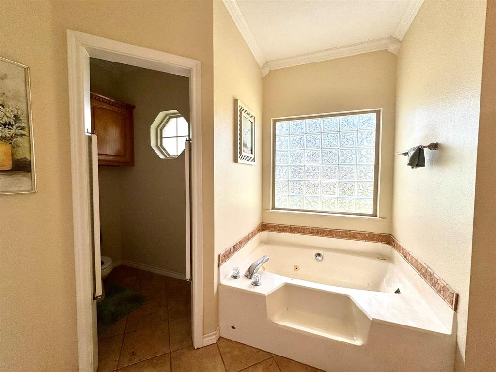 Corner jetted tub with tile surround, glass block window, crown molding, and tiled flooring