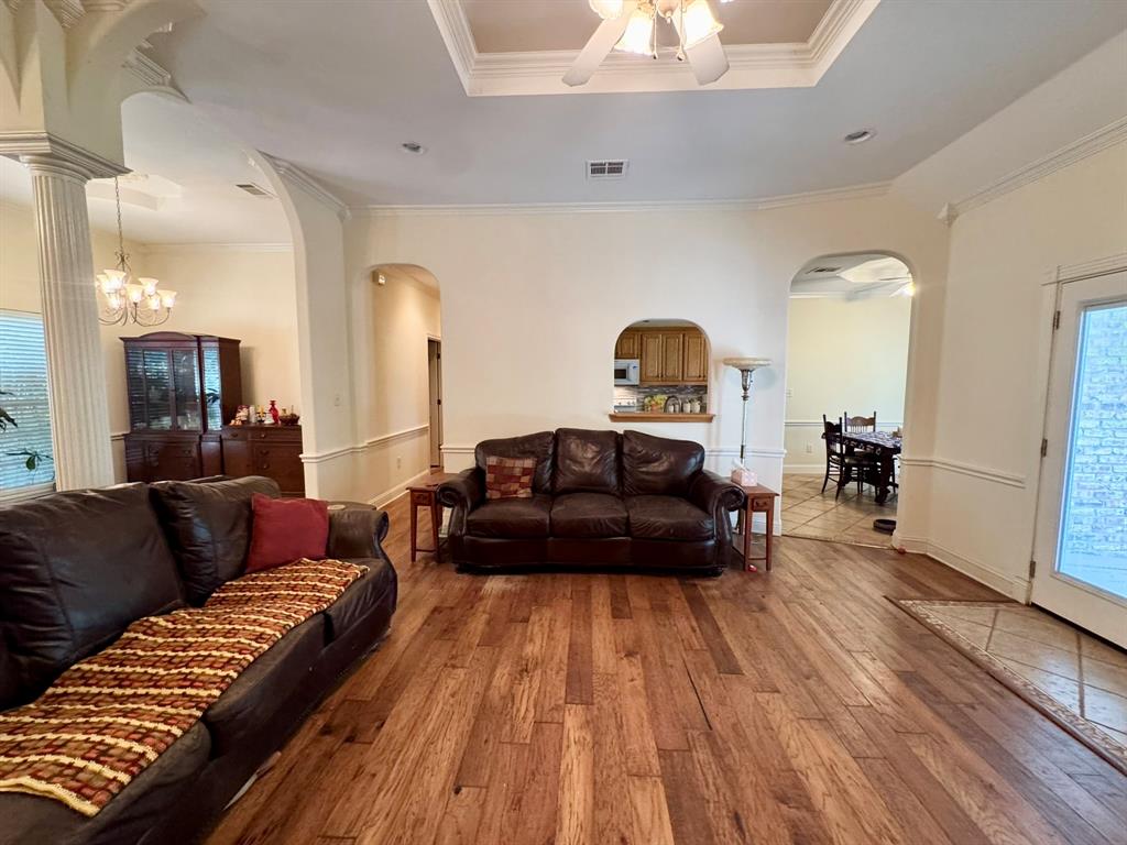 Open-concept interior featuring wood-finish flooring, tray ceilings with crown molding, decorative columns, and arched wall openings