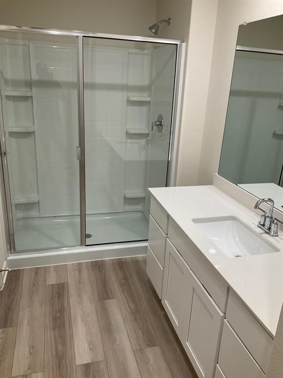 Bathroom featuring wood-finish flooring, white vanity cabinetry, a rectangular undermount sink with a quartz countertop, a large wall-mounted mirror, and a walk-in shower with a sliding glass door and integrated shelving