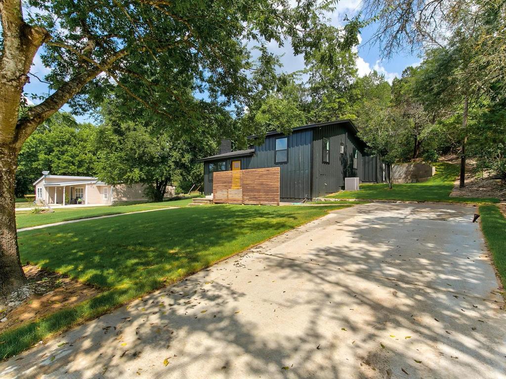 Exterior featuring black vertical siding, natural wood privacy screen, concrete driveway, and expansive lawn with mature trees