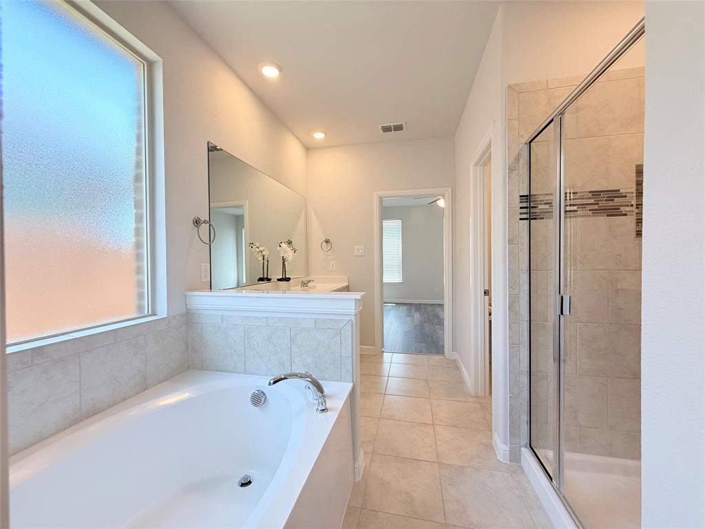 Soaking tub with chrome faucet, walk-in glass-enclosed shower featuring decorative tile inlay, ceramic tile flooring, and a large vanity mirror