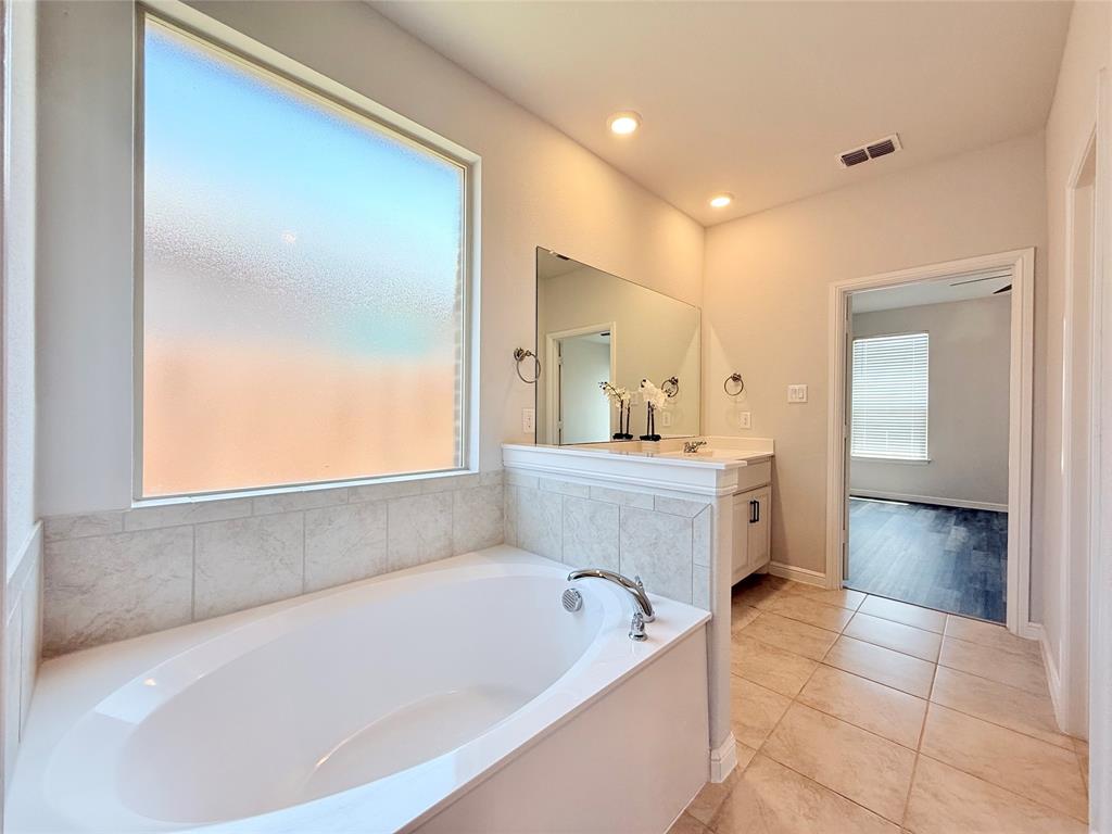 Oval soaking tub with tiled surround, obscured-glass window, large wall-mounted mirror, vanity cabinet, and tile flooring