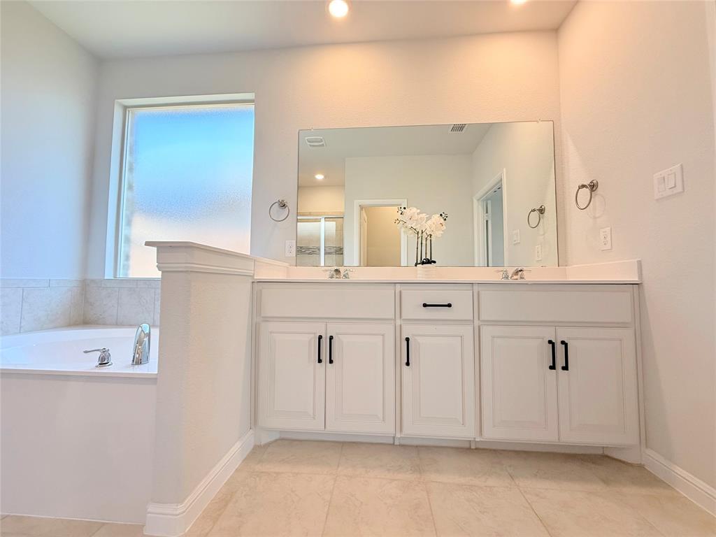 Bathroom featuring a soaking tub with tile surround, white cabinetry, dual-sink vanity, large rectangular mirror, and matte black hardware