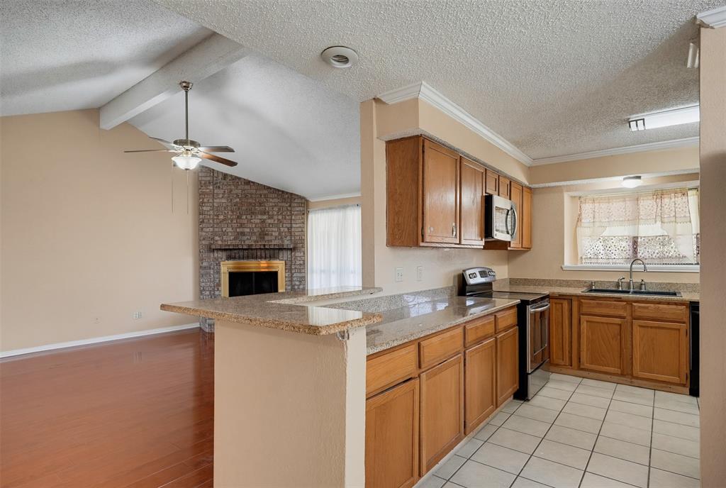 Kitchen featuring wood cabinetry, stone-finish countertops, tile flooring, and stainless steel appliances, with a vaulted ceiling and brick fireplace beyond the breakfast bar