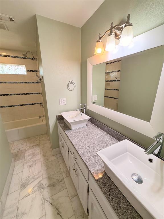 Double vanity featuring white rectangular vessel sinks, speckled stone countertops, and polished chrome faucets, complemented by white cabinetry, marble-style tiled flooring, and a shower-tub combination with decorative mosaic tile accents