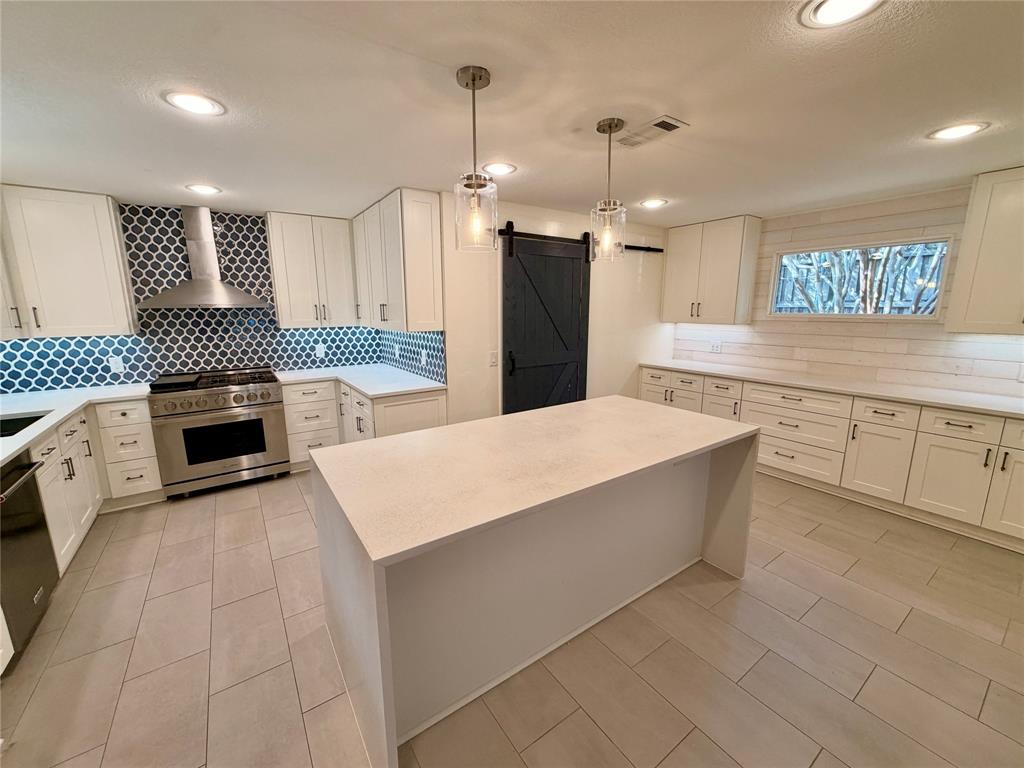 Kitchen featuring white cabinetry, a center island with a solid surface countertop, a patterned tile backsplash, stainless steel range with chimney-style hood, and a dark sliding barn-style door