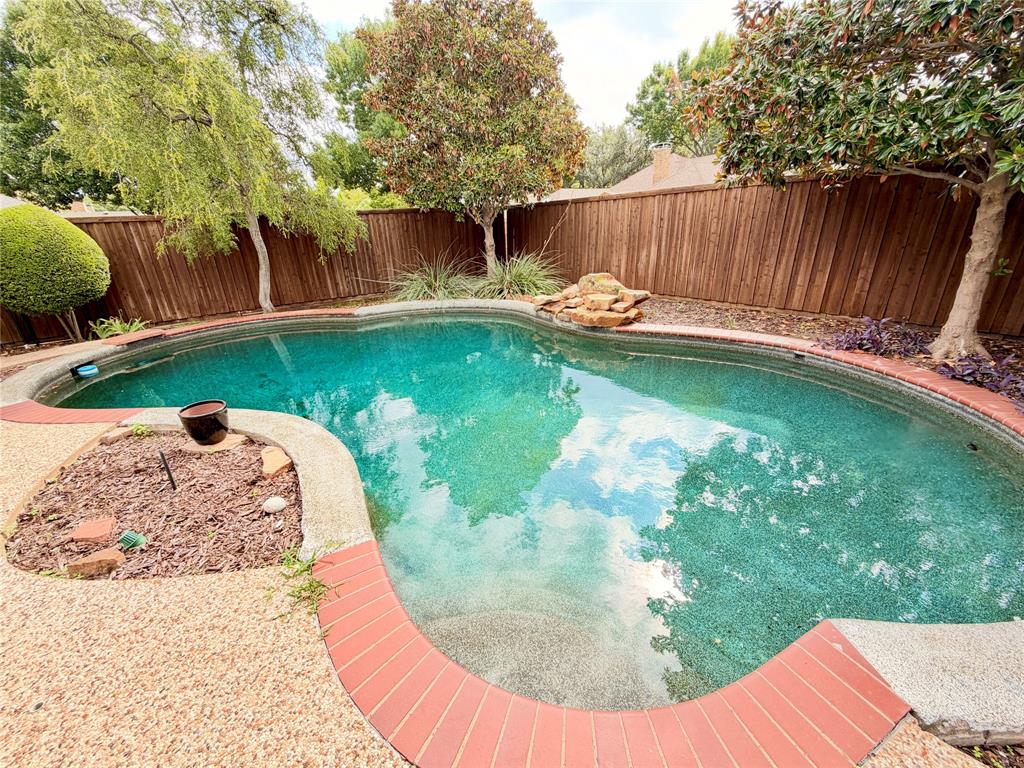 Curved swimming pool featuring a decorative rock water feature, red brick coping, textured concrete pool deck, and perimeter wood fencing