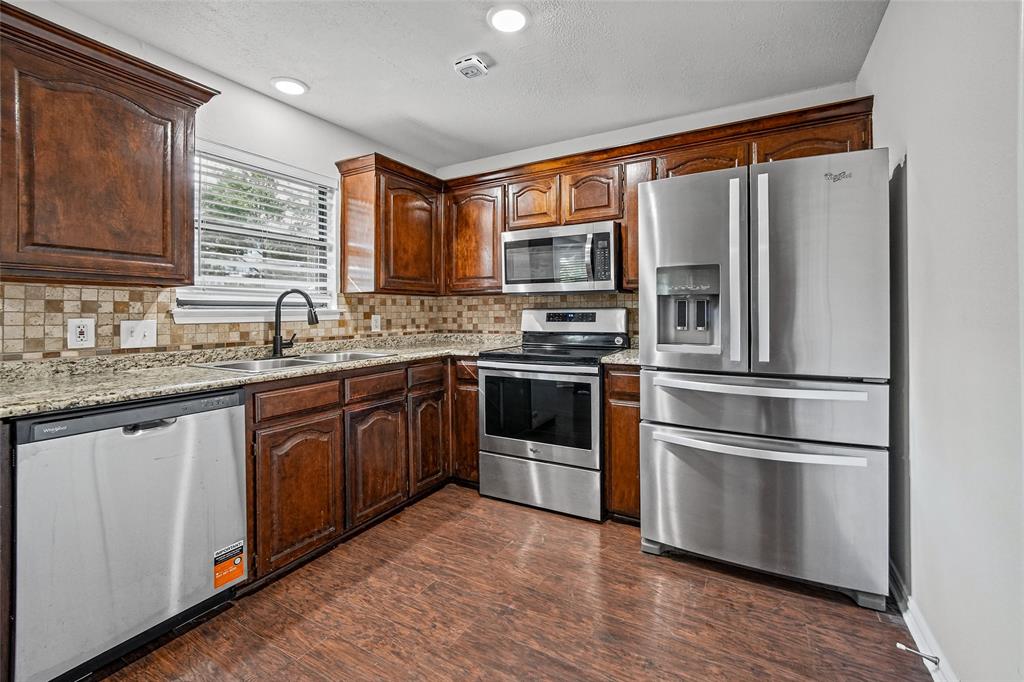 Kitchen featuring dark wood cabinetry, stone-surface countertops, a tiled backsplash, wood-finish flooring, and stainless steel appliances