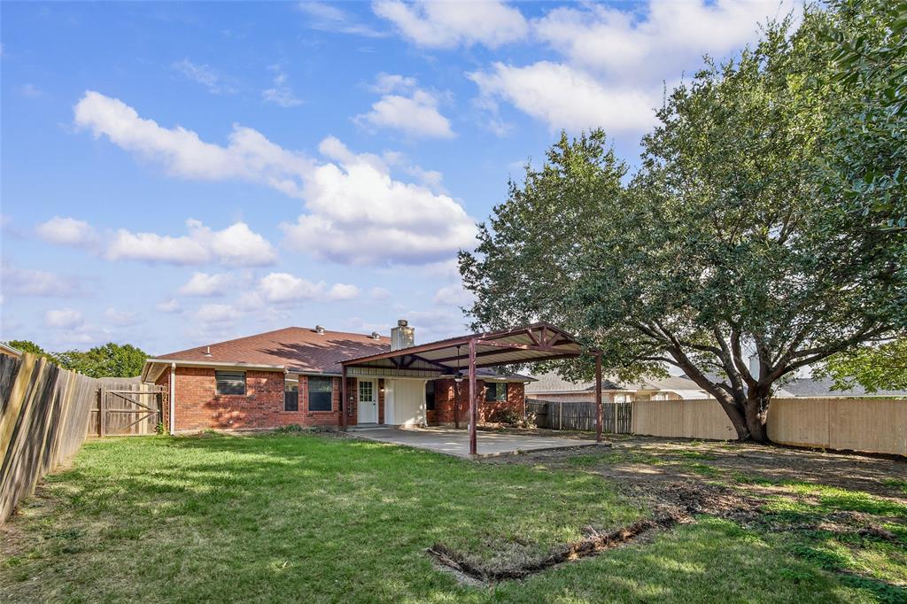 Rear exterior featuring a brick facade, covered patio area with metal supports, fenced yard, and a mature tree