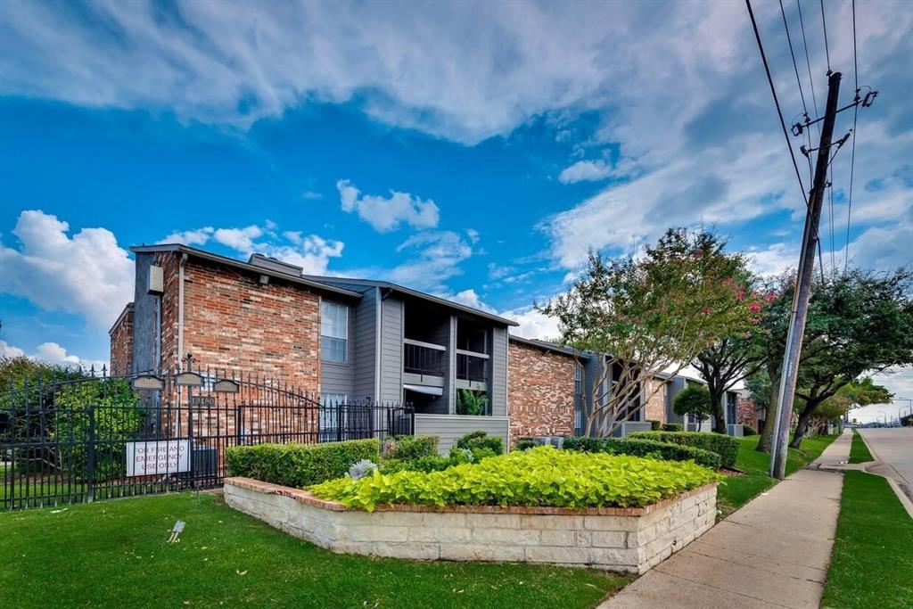 Multi-story building exterior featuring red brick and dark-toned paneling, raised stone-block landscaping, wrought-iron perimeter fencing, and mature trees Multi-story building exterior featuring red brick and dark-toned paneling, raised stone-block landscaping, wrought-iron perimeter fencing, and mature trees