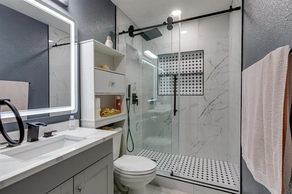 Bathroom featuring a vanity with a white countertop, illuminated wall mirror, glass-enclosed shower with tiled surround, built-in shower niche, and matte black plumbing fixtures