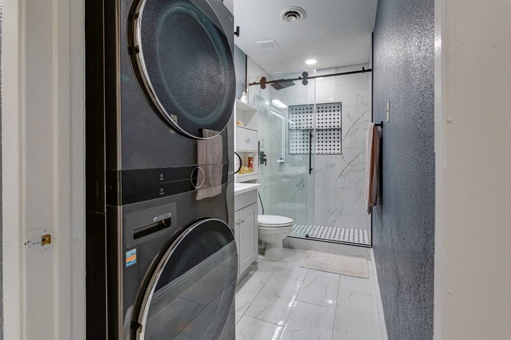 Integrated laundry area with stacked washer and dryer, bathroom featuring a walk-in shower with glass enclosure, large-format tile flooring, and matte black hardware